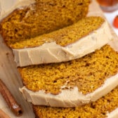 Three slices of pumpkin bread with light brown frosting on a wooden cutting board, with cinnamon sticks and a small orange candy pumpkin in the background.
