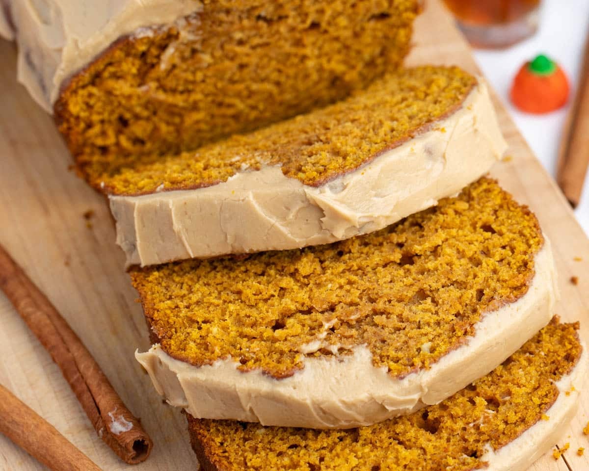 Three slices of pumpkin bread with light brown frosting on a wooden cutting board, with cinnamon sticks and a small orange candy pumpkin in the background.