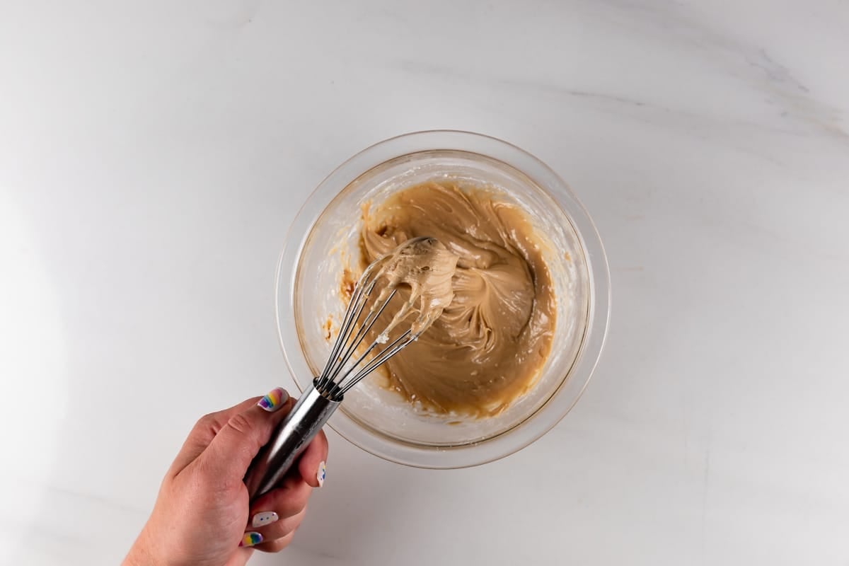 A hand holding a whisk mixes light brown batter in a glass bowl on a white countertop. The persons nails are painted with colorful designs.