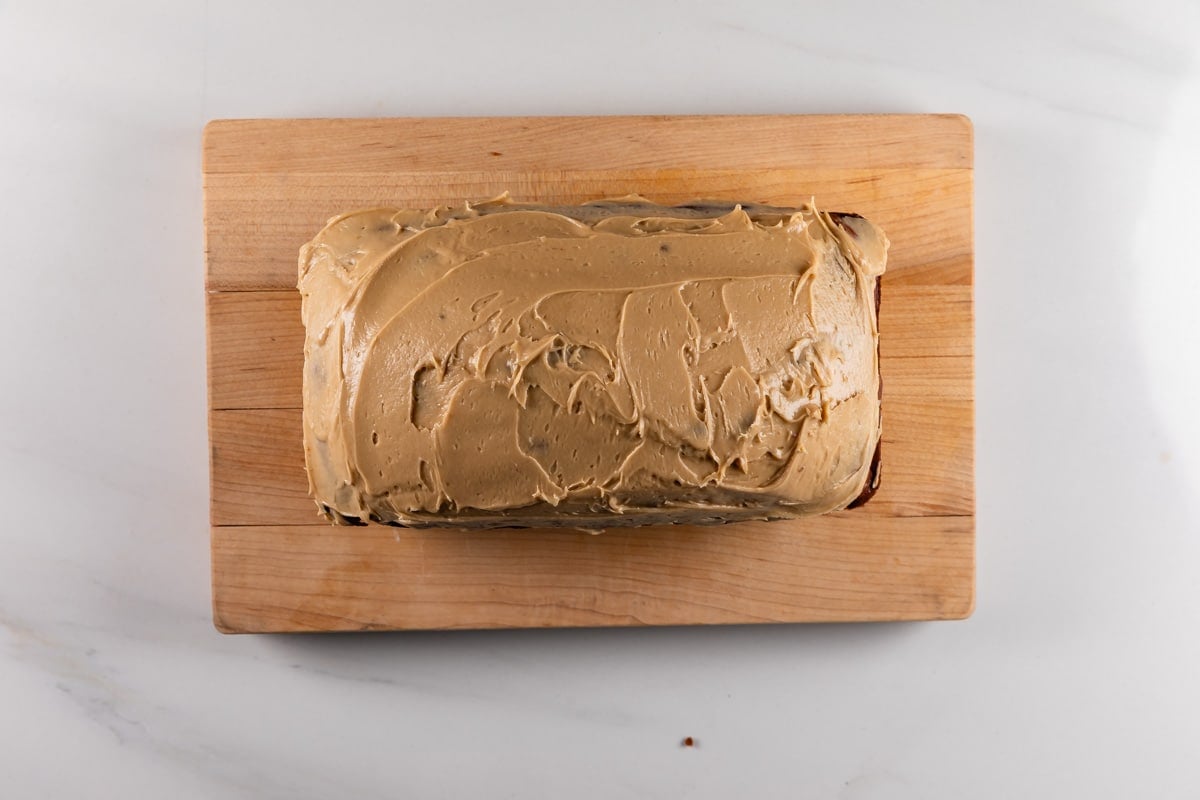 A rectangular loaf cake with light brown frosting sits on a wooden cutting board, viewed from above, on a white countertop.