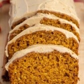 Sliced pumpkin bread with light brown frosting on a wooden board, with a cinnamon stick beside it and a colorful cloth in the background.