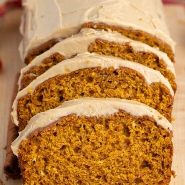 Sliced pumpkin bread with light brown frosting on a wooden board, with a cinnamon stick beside it and a colorful cloth in the background.