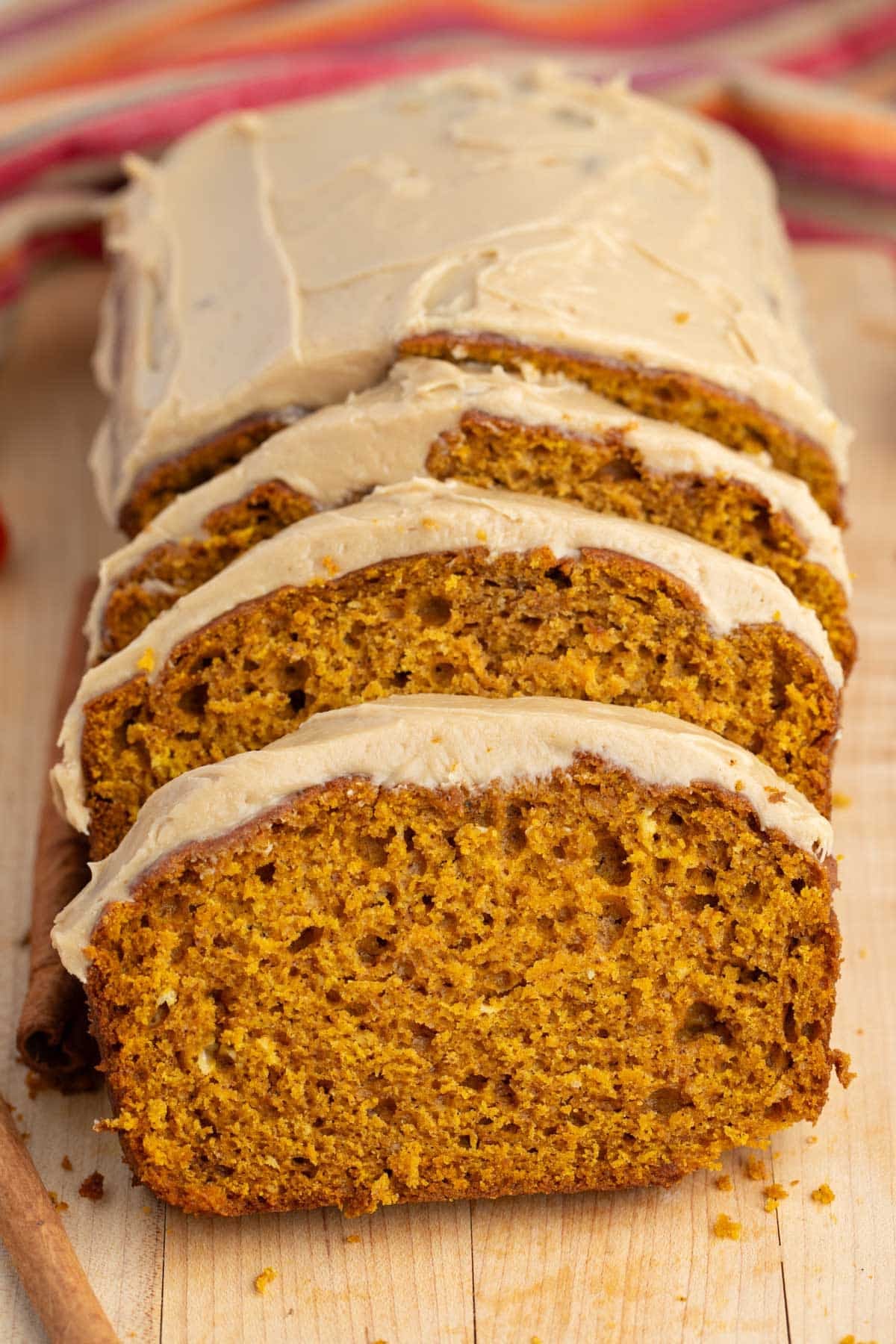 Sliced pumpkin bread with light brown frosting on a wooden board, with a cinnamon stick beside it and a colorful cloth in the background.