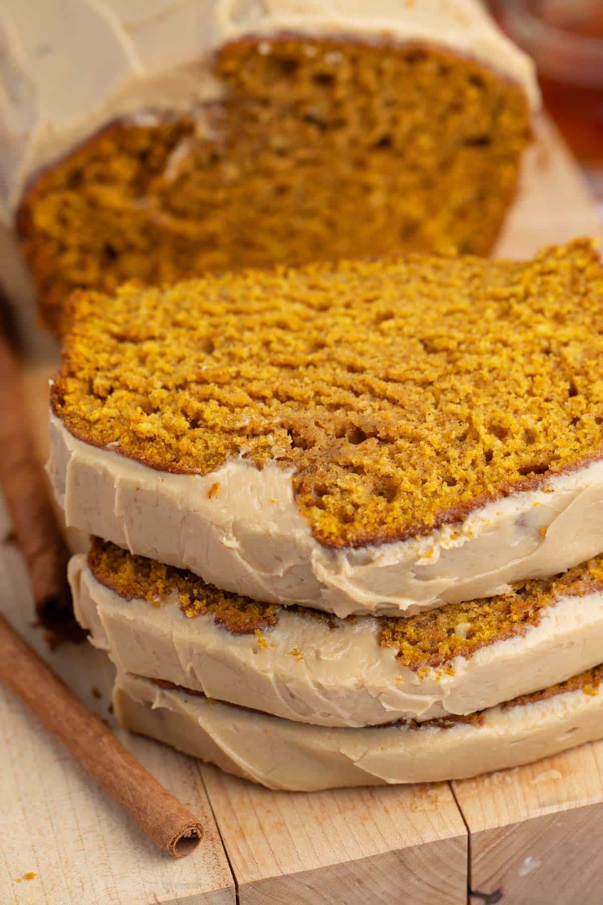 Two thick slices of pumpkin bread with light brown icing stacked on a wooden cutting board. A cinnamon stick lies next to the bread, and more of the loaf is visible in the background.