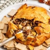 A wedge of baked brie topped with pecans and apple pieces inside a flaky pastry crust, surrounded by square crackers on a plate.