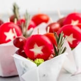 Small parchment cups containing red wax-covered cheeses cut with star shapes, grapes, and rosemary sprigs.
