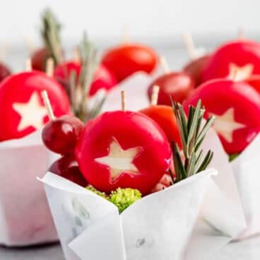 Small parchment cups containing red wax-covered cheeses cut with star shapes, grapes, and rosemary sprigs.