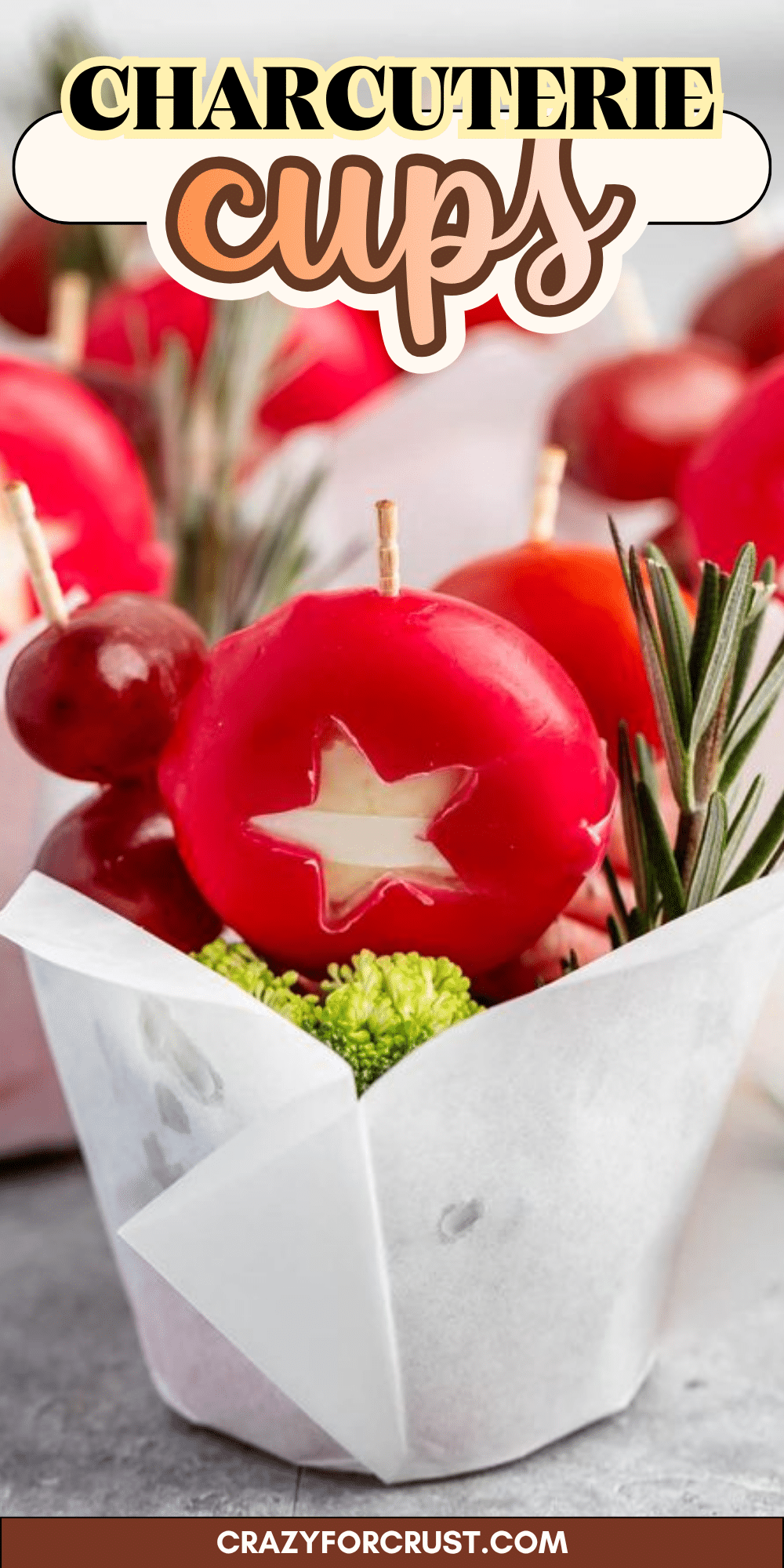 A charcuterie cup featuring a wax-covered cheese wheel with a star-shaped cutout, grapes, and rosemary sprigs in a white paper container.