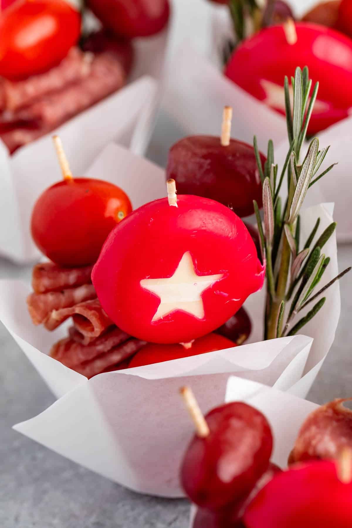 Individual appetizer cone containing a cherry tomato, folded salami, a mini cheese wheel with a star cutout, and a sprig of rosemary on a toothpick.
