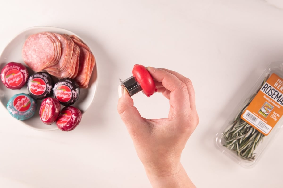 A hand holds a Babybel cheese wheel with a red wax coating, with a plate of sliced salami and Babybel cheeses and a package of rosemary nearby.