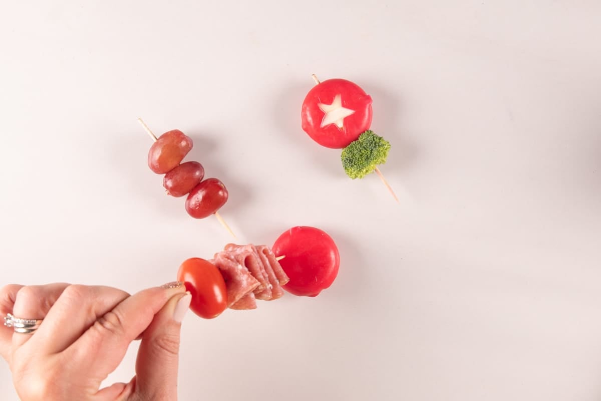 A hand holds a skewer with a cherry tomato and folded deli meat, next to two other skewers featuring grapes, broccoli, and round red cheeses, one with a star cutout.