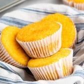 Four golden-yellow corn muffins in paper liners nestled in a blue and white striped cloth.