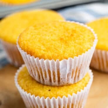 Two corn muffins in white paper liners stacked on a wooden board, with more muffins in a tin in the background.