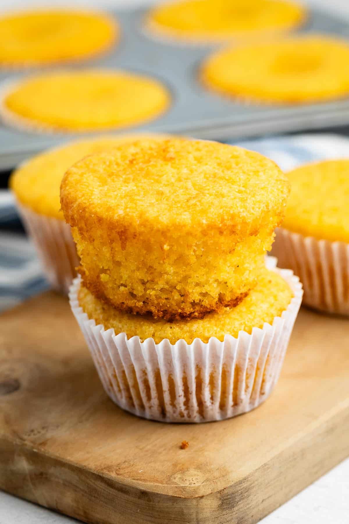 A stack of two cornbread muffins in white paper liners on a wooden board, with more muffins in a tin in the background.