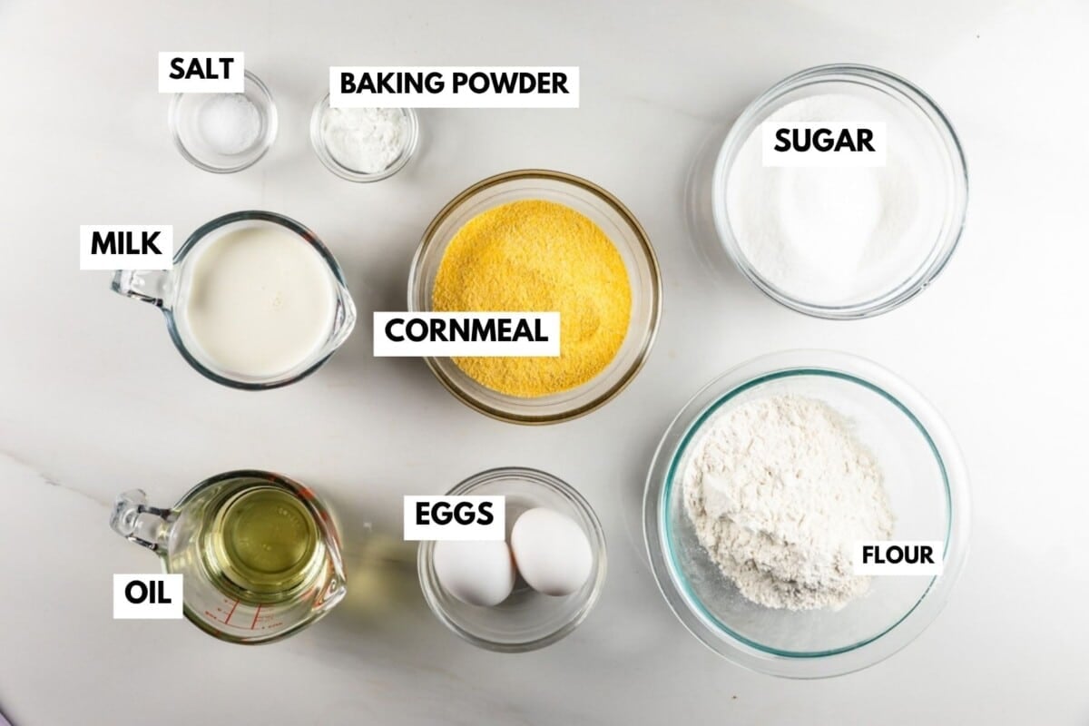 Top-down view of baking ingredients in bowls labeled salt, baking powder, sugar, milk, cornmeal, oil, eggs, and flour.