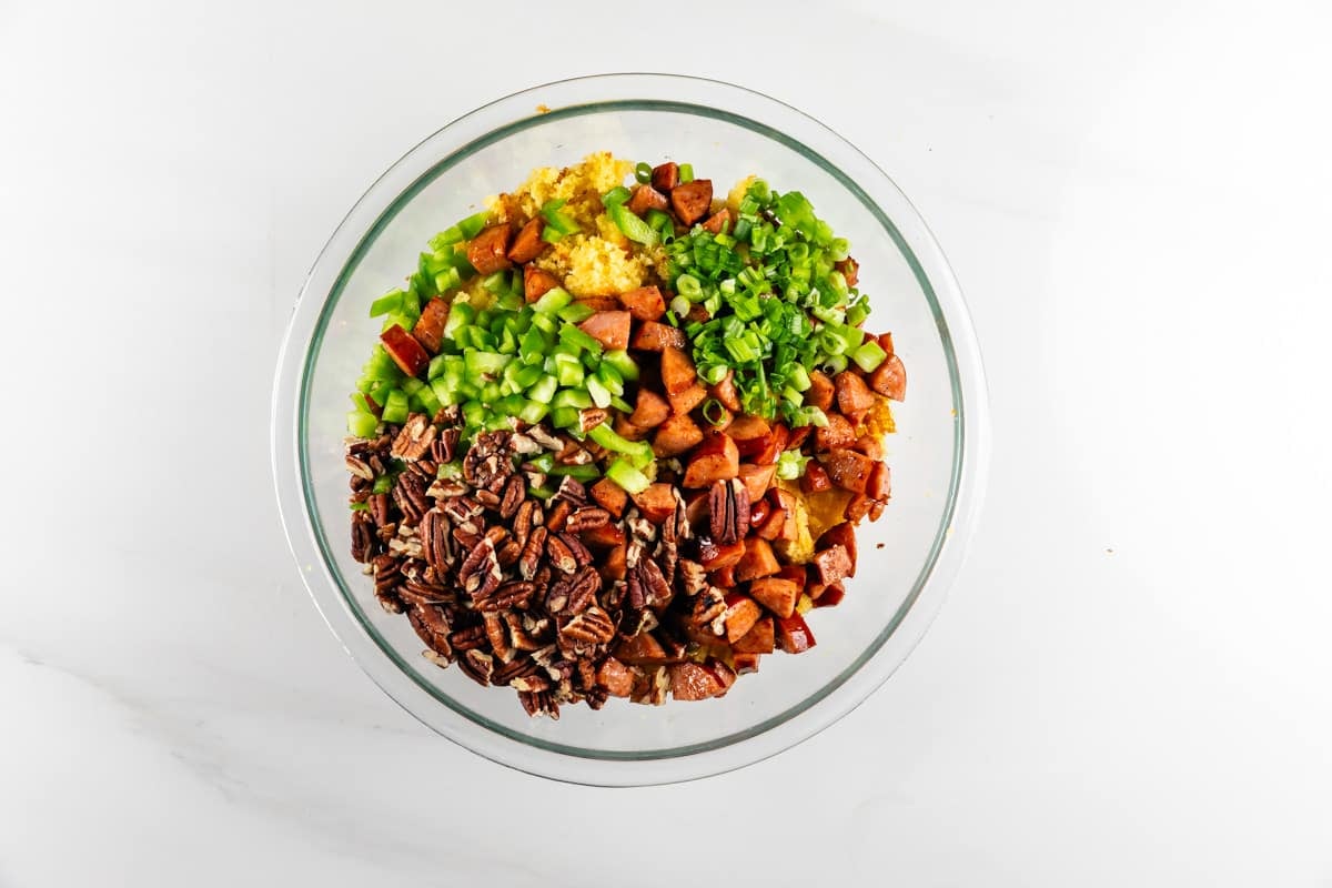 A glass bowl filled with sections of cornbread, diced green bell peppers, sliced sausage, green onions, and pecans.