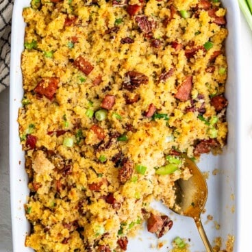 A rectangular white baking dish filled with cornbread dressing containing bits of sausage, pecans, and green onions, with a gold serving spoon resting inside.