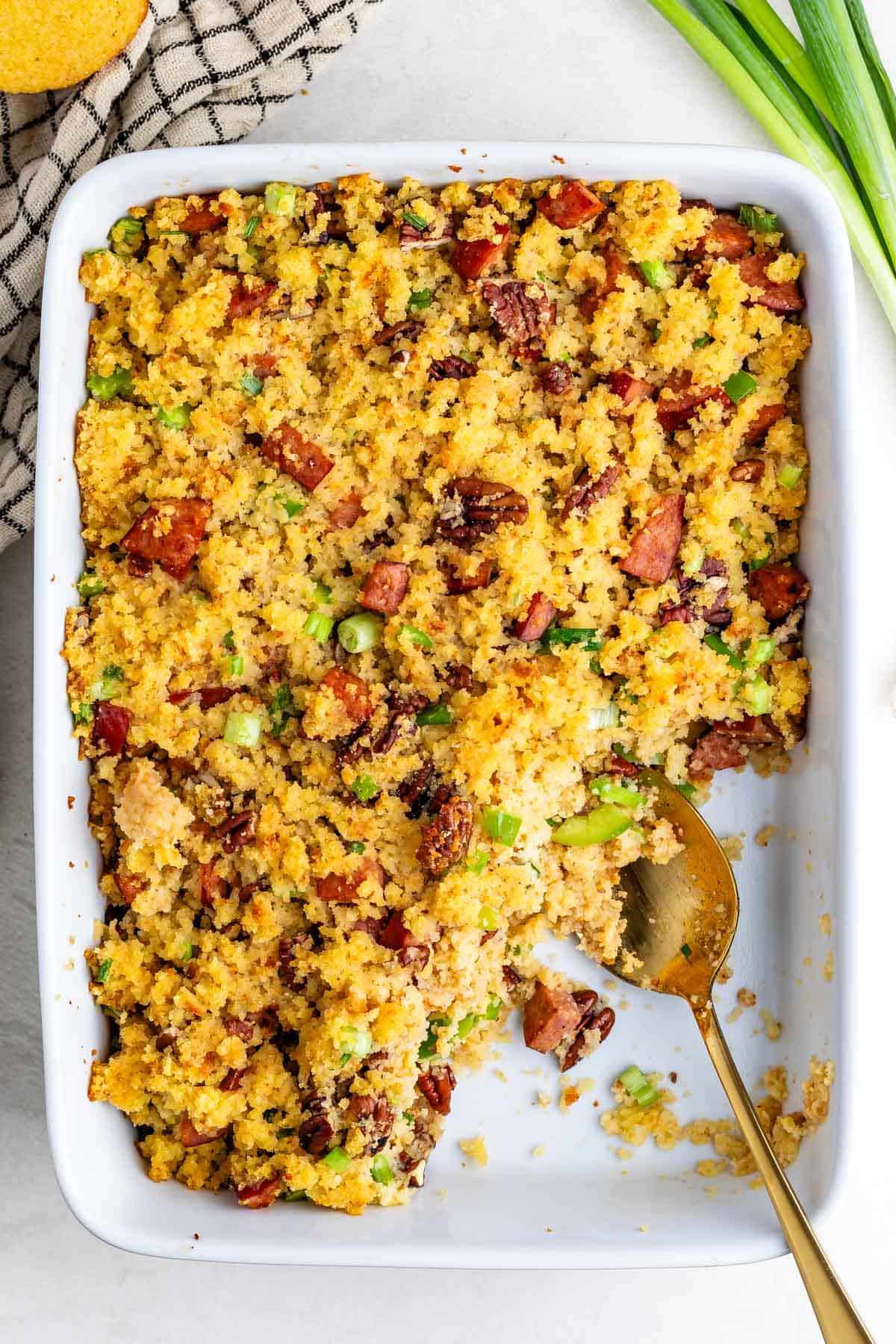 A rectangular white baking dish filled with cornbread dressing containing bits of sausage, pecans, and green onions, with a gold serving spoon resting inside.