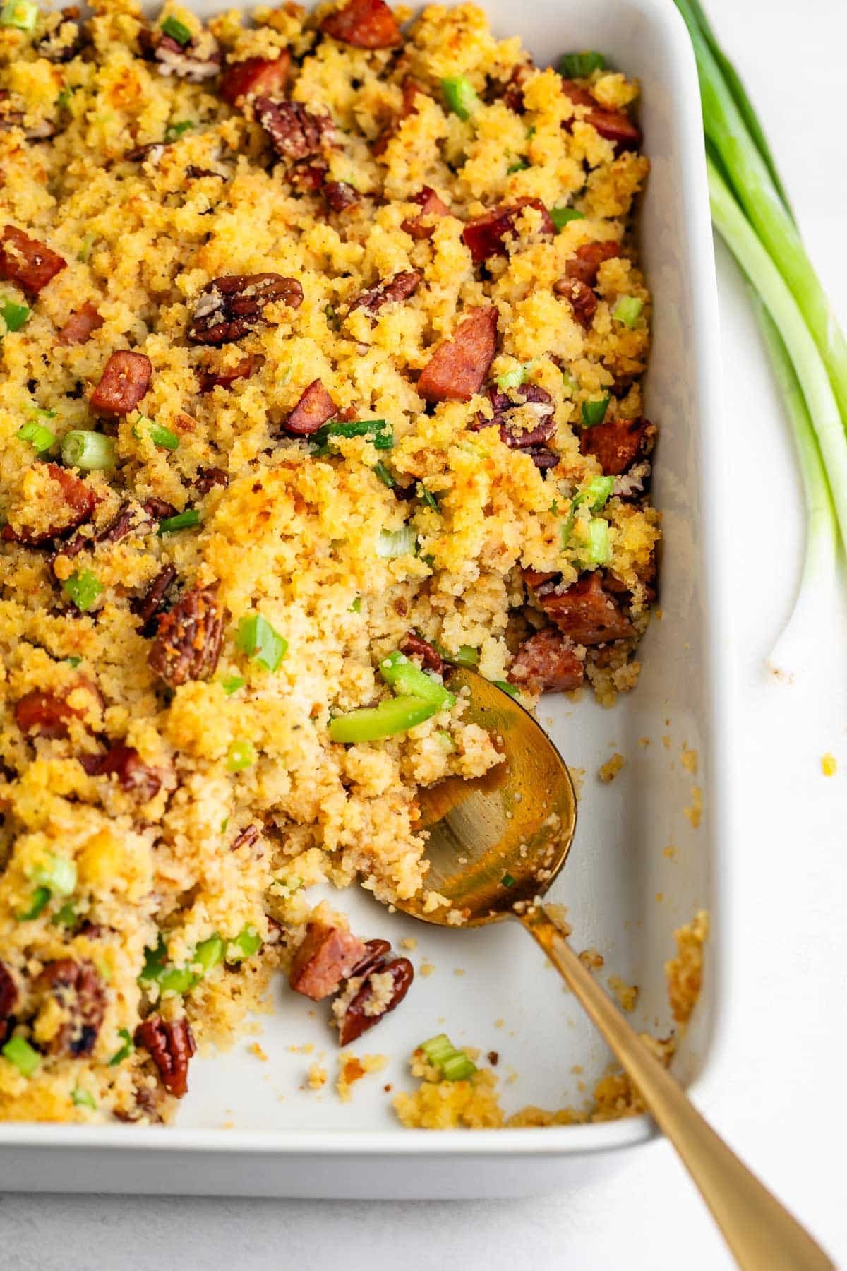 Cornbread dressing with sausage, pecans, and green onions in a white baking dish with a gold serving spoon.
