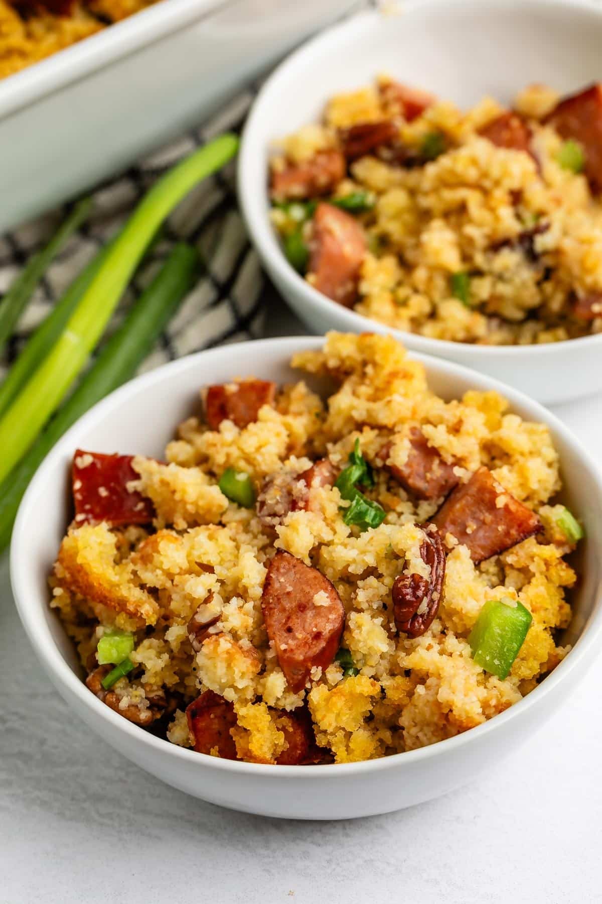 Two white bowls filled with cornbread dressing containing sliced sausage, pecans, and green bell pepper.