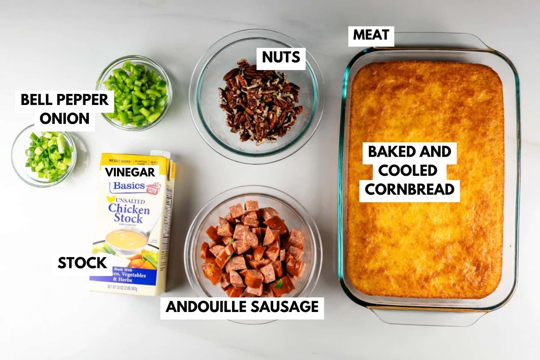 Ingredients for a recipe arranged on a counter: chopped bell pepper, chopped onion, pecans, a carton of chicken stock, sliced andouille sausage, and a rectangular pan of baked cornbread.