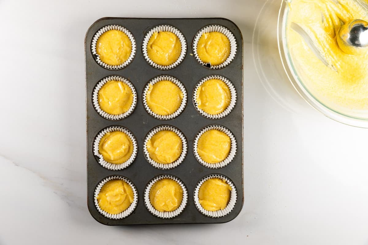 A metal muffin pan filled with twelve paper liners containing yellow cake batter, next to a glass bowl of batter and a whisk.