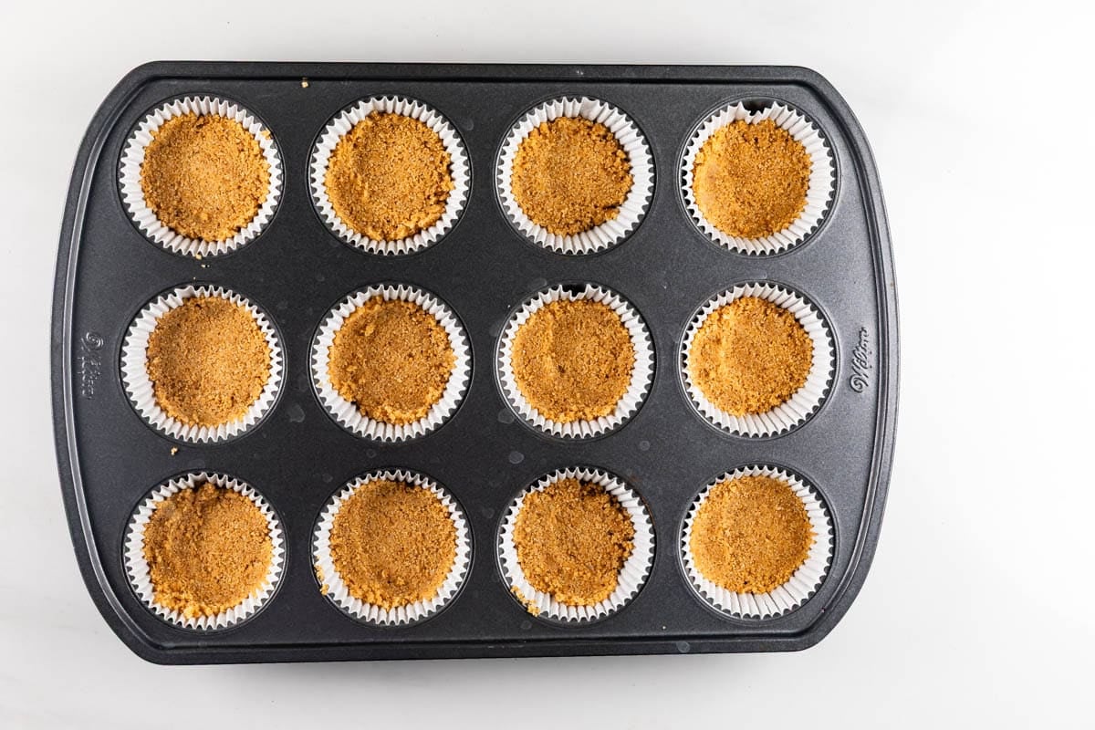 A muffin tin containing twelve paper liners filled with pressed graham cracker crusts.