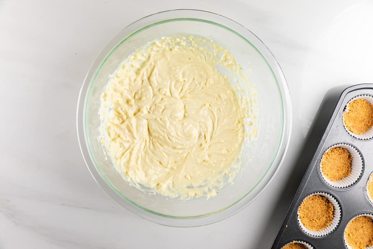 A glass bowl of creamy pale batter next to a muffin tin lined with paper cups and filled with cookie crumb crusts.