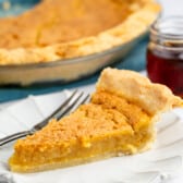 A slice of golden yellow chess pie on a white scalloped plate with a fork, with the remaining pie in a glass dish in the background.