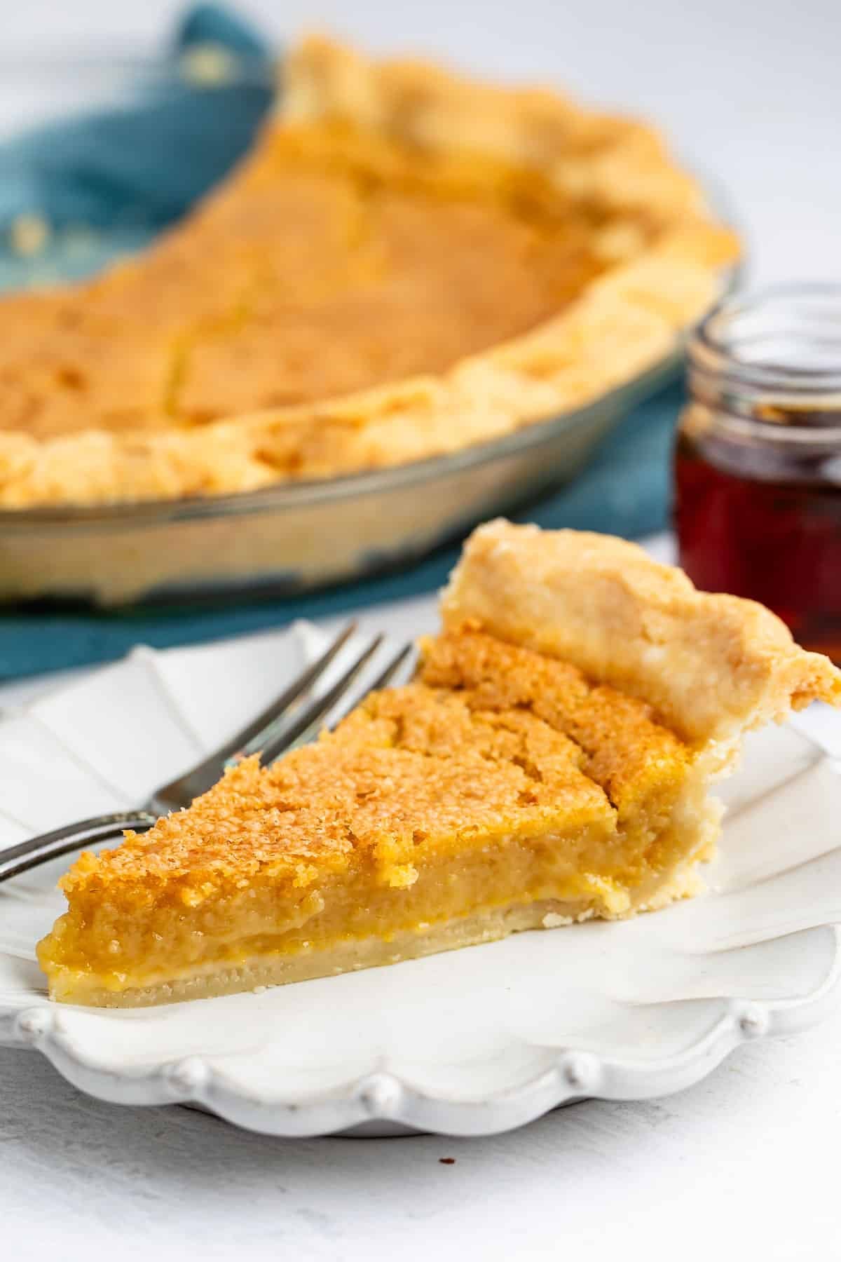 A slice of golden yellow chess pie on a white scalloped plate with a fork, with the remaining pie in a glass dish in the background.