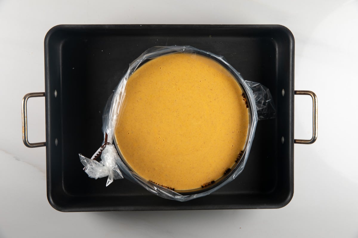 A pumpkin cheesecake in a springform pan wrapped in plastic, sitting inside a black roasting pan.