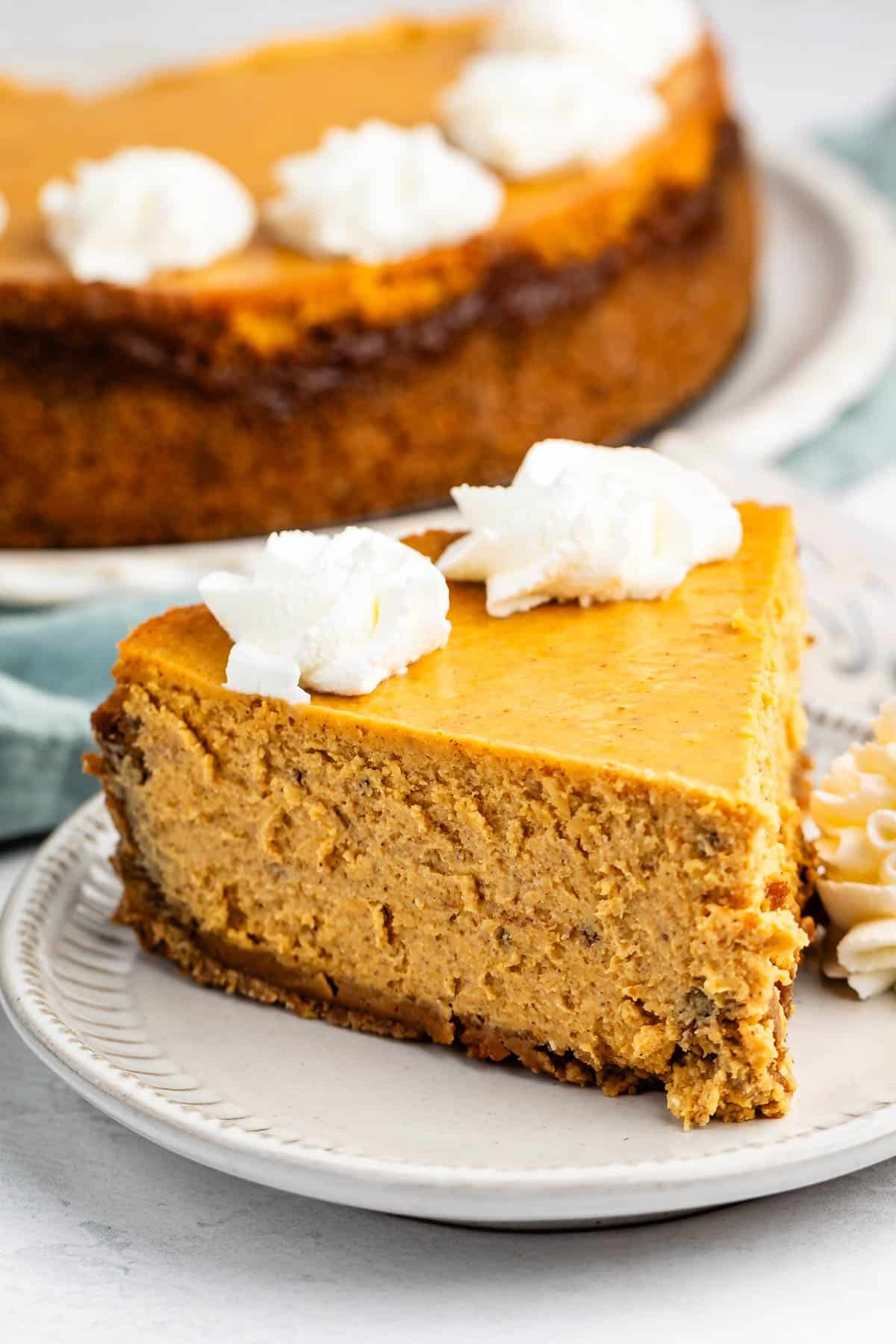 A slice of pumpkin cheesecake topped with whipped cream on a small plate, with the rest of the cheesecake visible in the background.